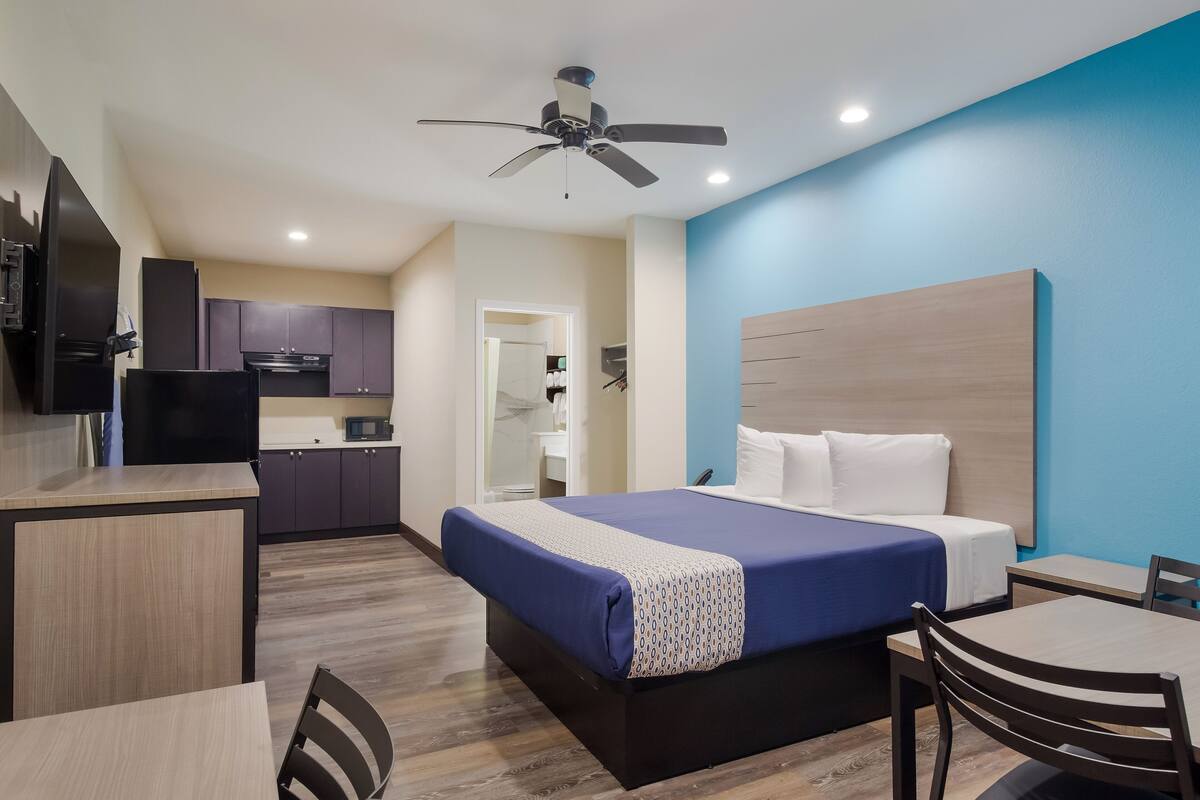 A hotel room with a bed, a desk, a chair, a kitchenette, and a ceiling fan.