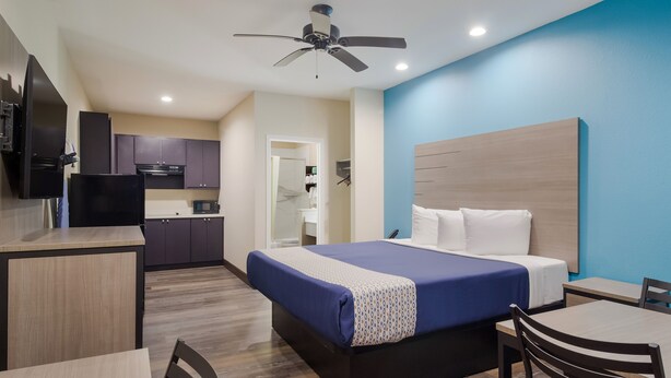Une chambre d’hôtel moderne équipée d’un lit, d’un bureau, d’une chaise, d’une kitchenette et d’un ventilateur de plafond.