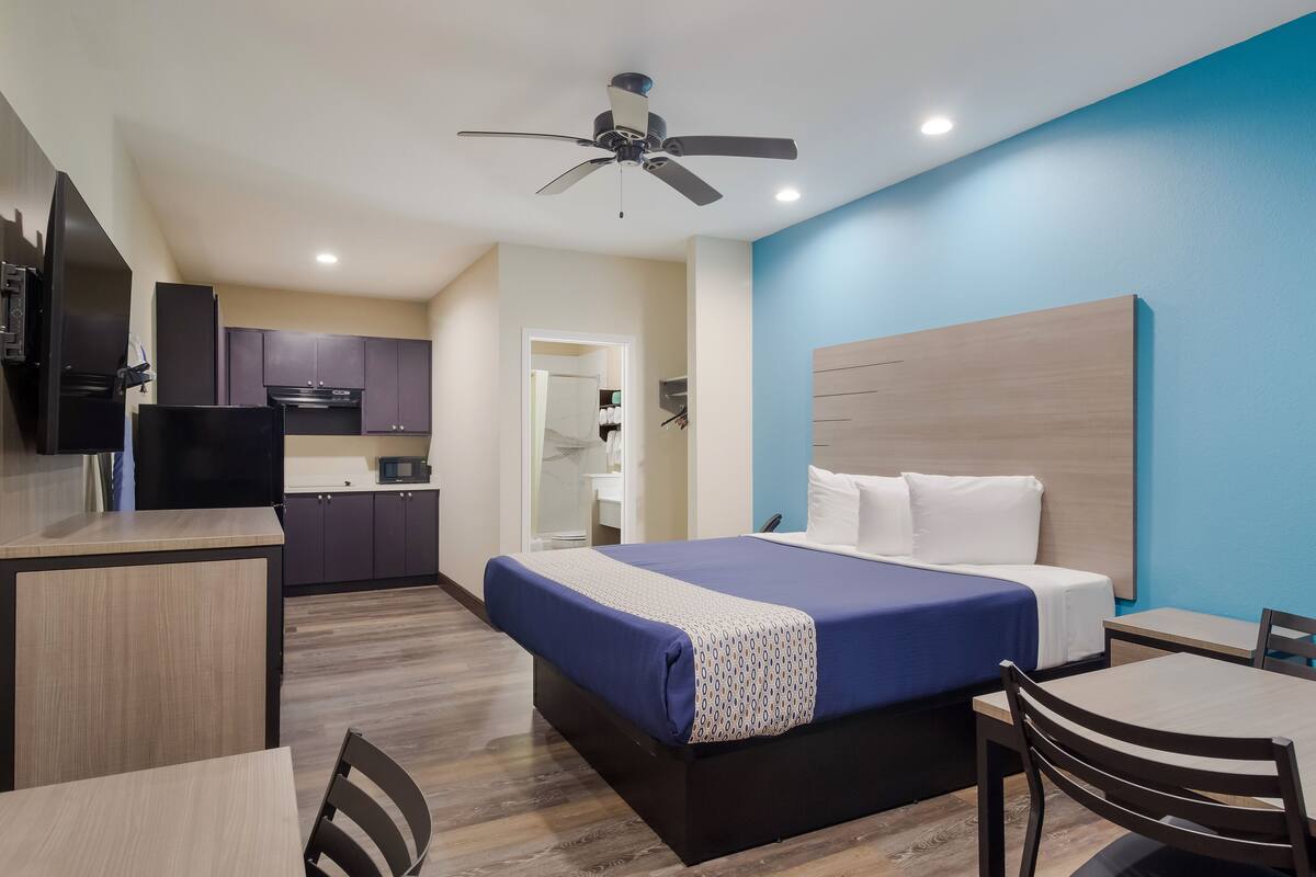 A modern hotel room with a bed, a desk, a chair, a kitchenette, and a ceiling fan.