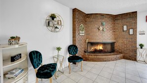 Een knusse woonkamer met een metselwerk open haard, twee comfortabele fauteuils, een witte boekenkast en een klein bijzettafeltje.
