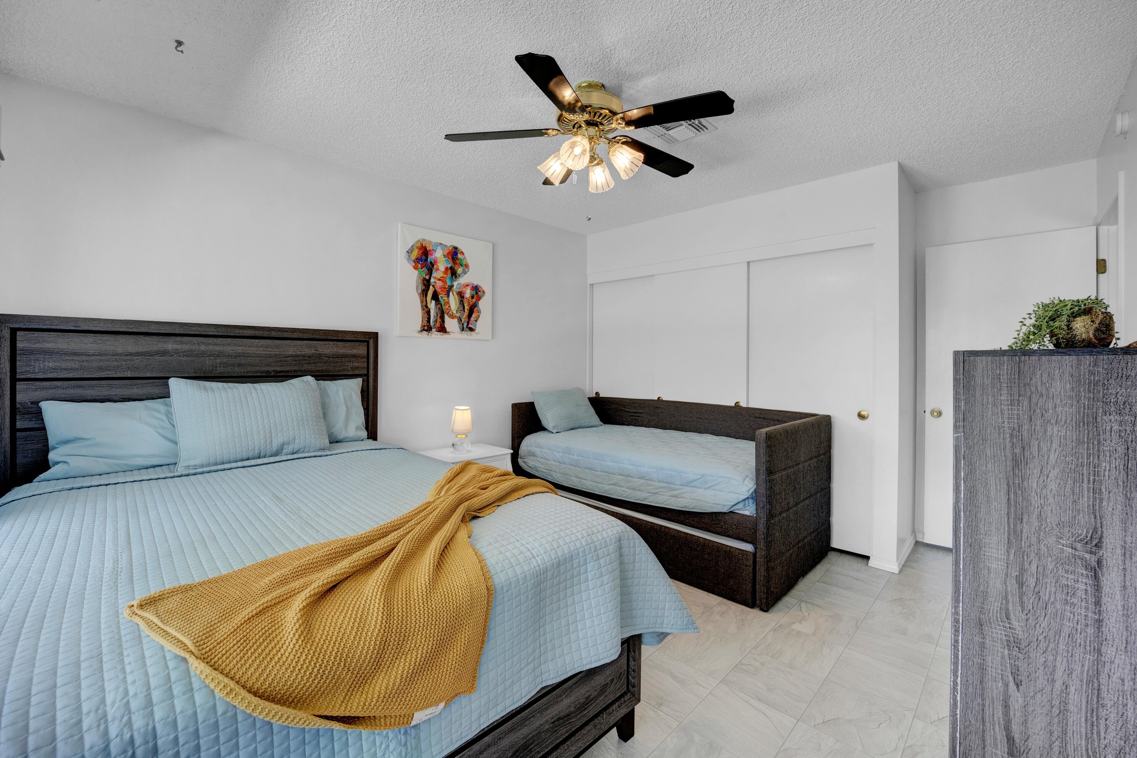 A bedroom with two beds, a ceiling fan, a nightstand with a lamp, and a door with a plant on top.