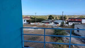 A view from a balcony with a clear sky, featuring a dirt road, several buildings, and a distant hill.
