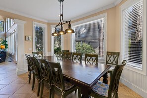 Une salle à manger avec une grande table en bois, entourée de chaises vertes, un lustre et de grandes fenêtres équipées de stores.