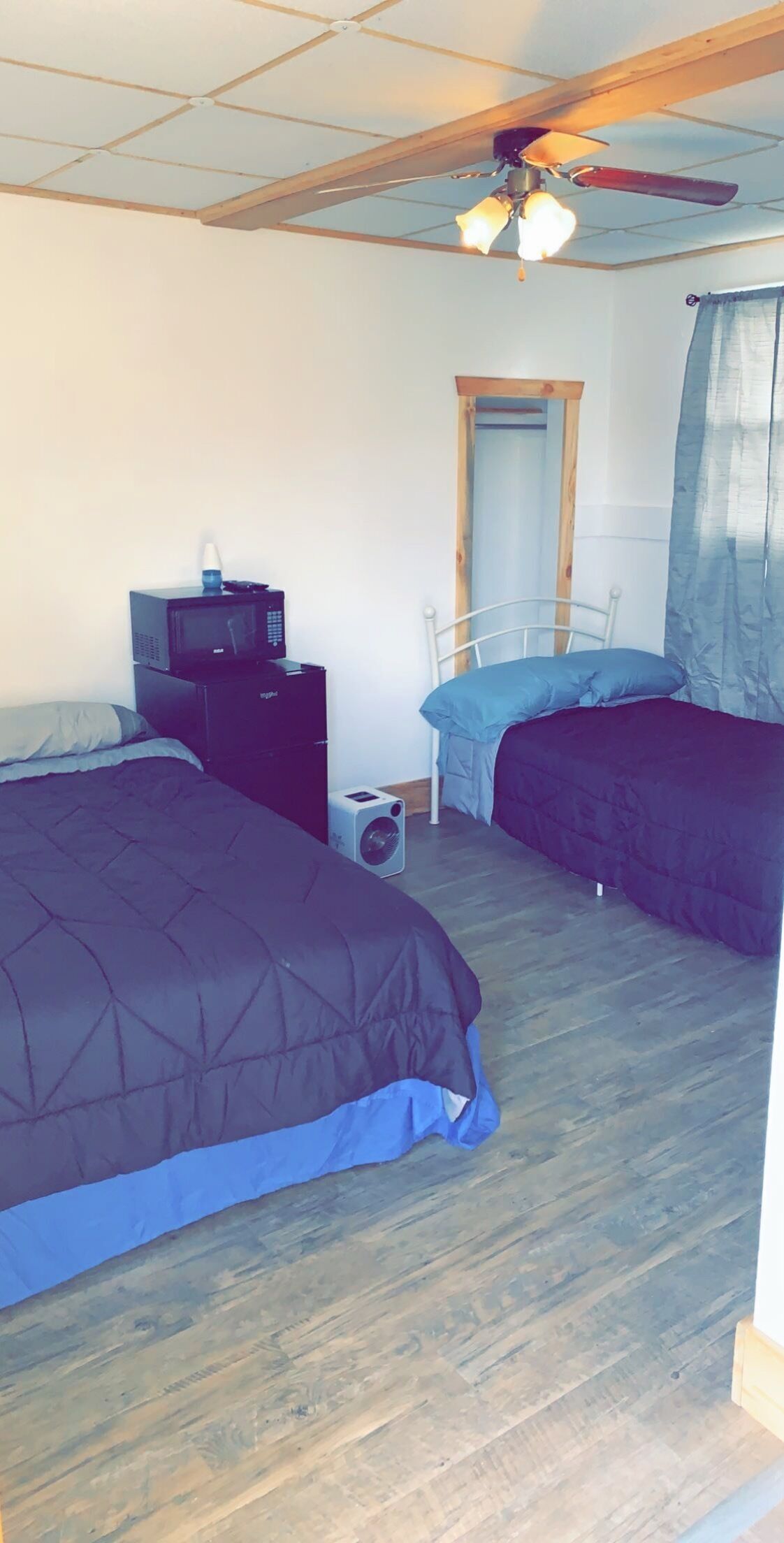 A bedroom with two beds, a ceiling fan, a heater, a mirror, and a window with curtains.
