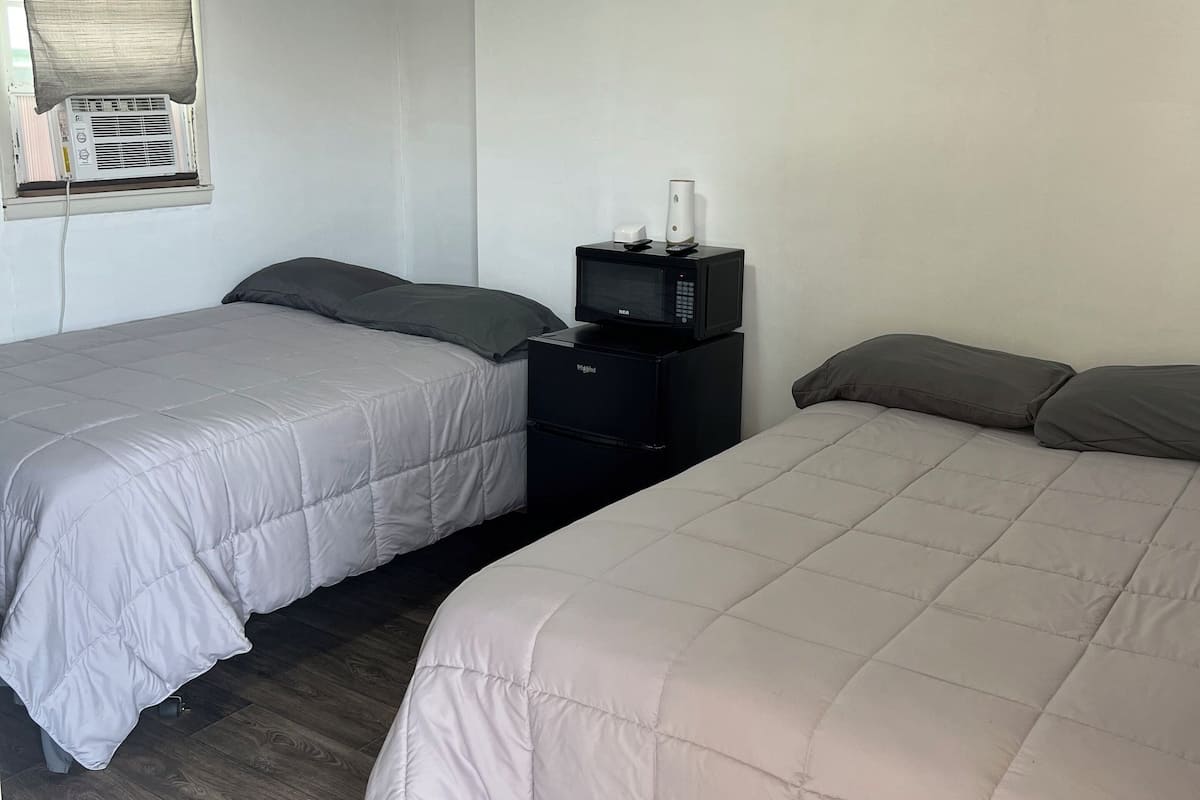 Quarto de hotel com duas camas, uma pequena mesa preta com micro-ondas, janela com persianas e uma luminária no teto.