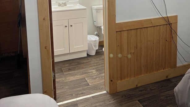 A room with a wooden-framed door leading to a bathroom, a mounted TV, and a visible light switch. - High Plains Inn (Newell)