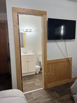 Una habitación con una puerta de madera que da a un baño, un televisor montado y un interruptor de luz a la vista.