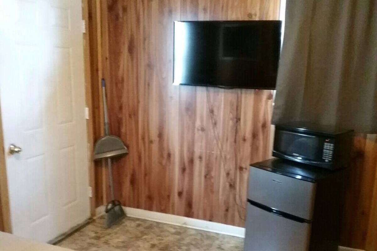 Quarto compacto com porta de madeira, televisão fixada na parede, micro-ondas em um pequeno armário e uma vassoura encostada na parede.