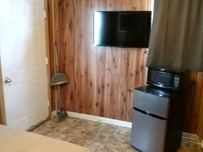 A compact room with a wooden door, a television mounted on the wall, a microwave on a small cabinet, and a broom leaning against the wall.