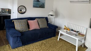 A living room with a dark blue sofa, patterned cushions, a round mirror, a painting of a pier, a white lamp, a white side table with fruits and snacks, and an air conditioning unit. - Lodge , air con , Free Parking , Mini bar (Hove)