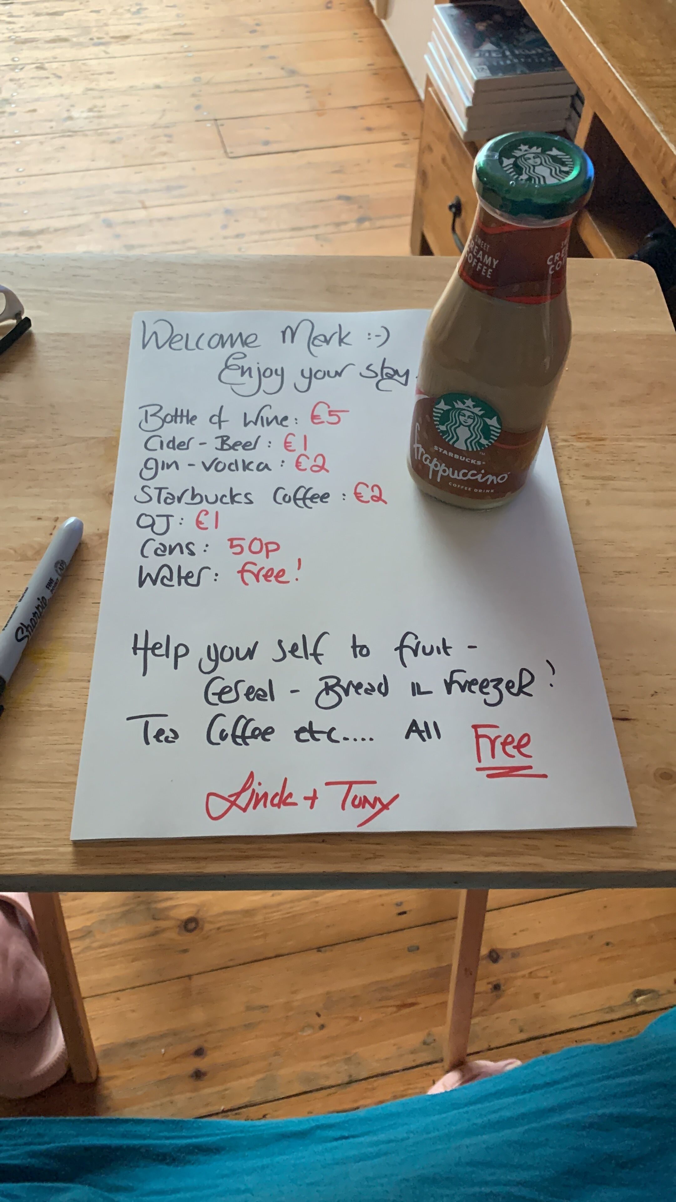 Ein handgeschriebenes Schild mit Preisen für verschiedene Artikel wie Wein, Apfelwein und Kaffee, mit einer Starbucks-Flasche auf einem Holztisch.