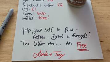 Ein handgeschriebenes Schild mit Preisen für verschiedene Artikel wie Wein, Apfelwein und Kaffee, mit einer Starbucks-Flasche auf einem Holztisch.
