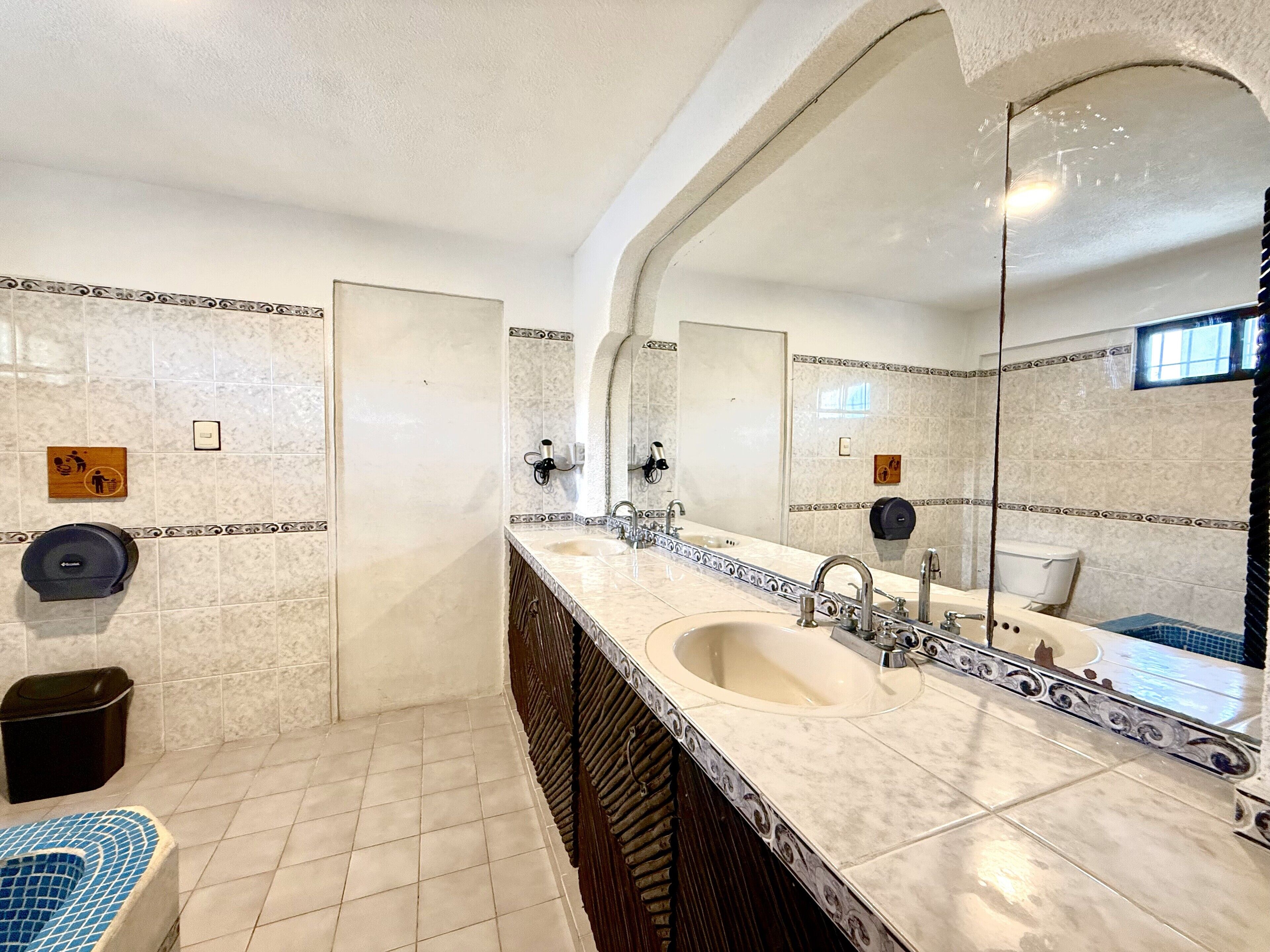 A bathroom with a large mirror, two sinks, a toilet, and a trash bin.