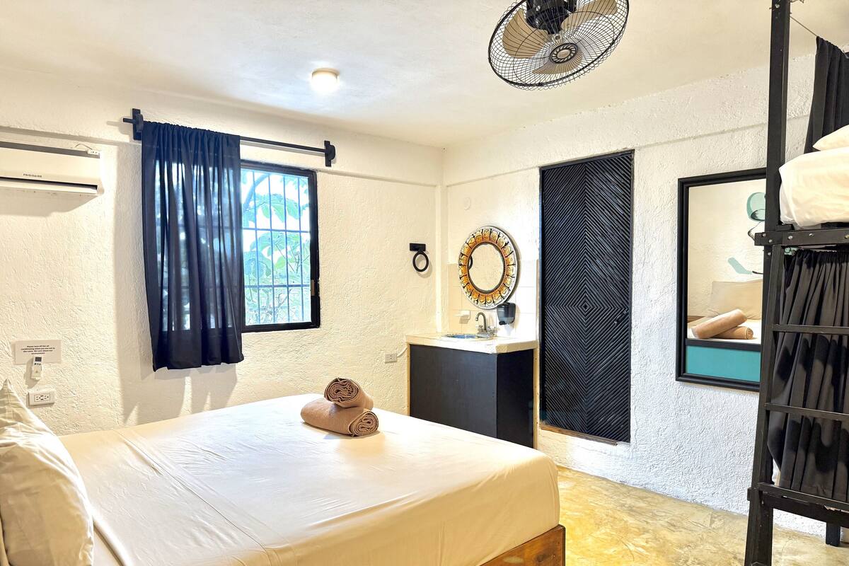 A bedroom with a bed, a window with curtains, a mirror, a sink, and a fan.