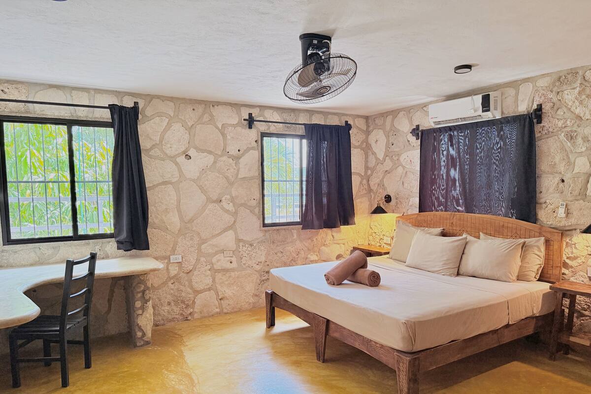 A room with a stone wall, a bed with a wooden headboard, a desk with a chair, and a window with curtains.