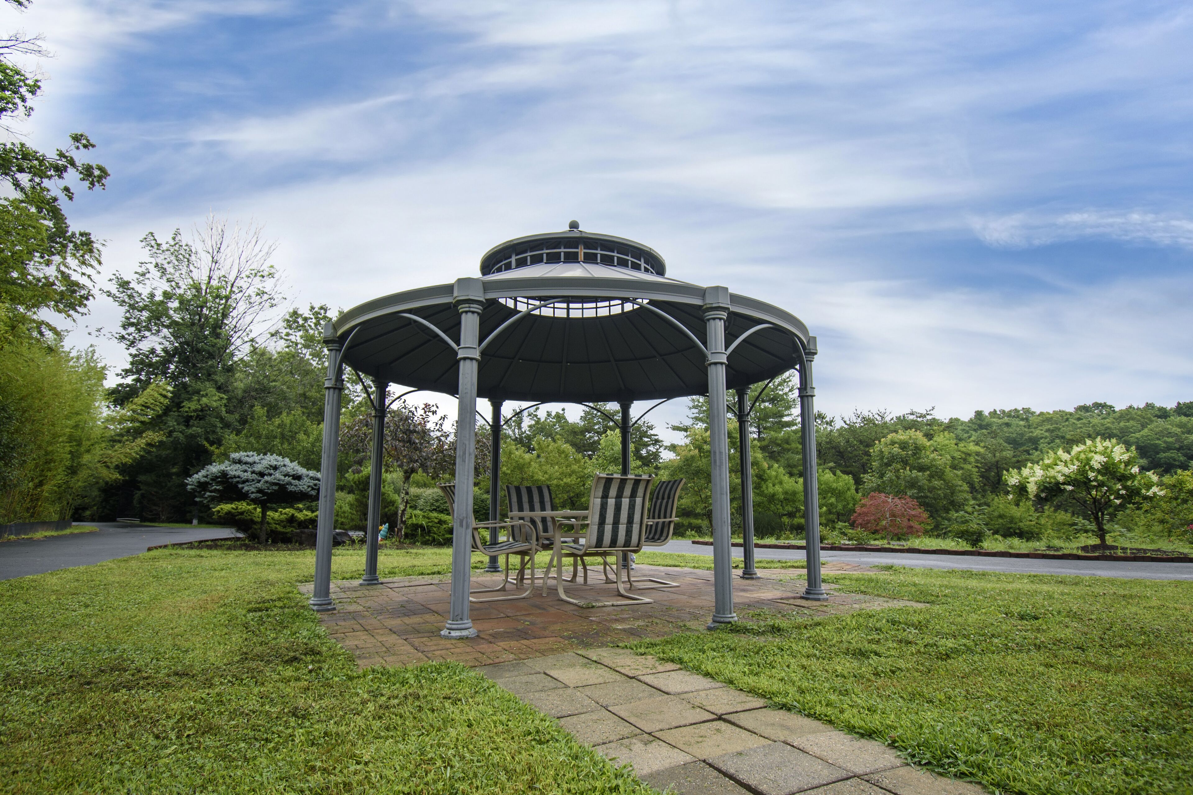Gazebo dal tetto circolare con quattro sedie sotto, circondato da erba e alberi.