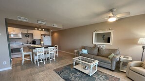 Een woonkamer met een bank, salontafel en een eetgedeelte met stoelen, en een keuken op de achtergrond.