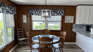 Un comedor con una mesa redonda y seis sillas de madera, una lámpara de araña, y una cocina con armarios blancos y salpicadero de azulejos.