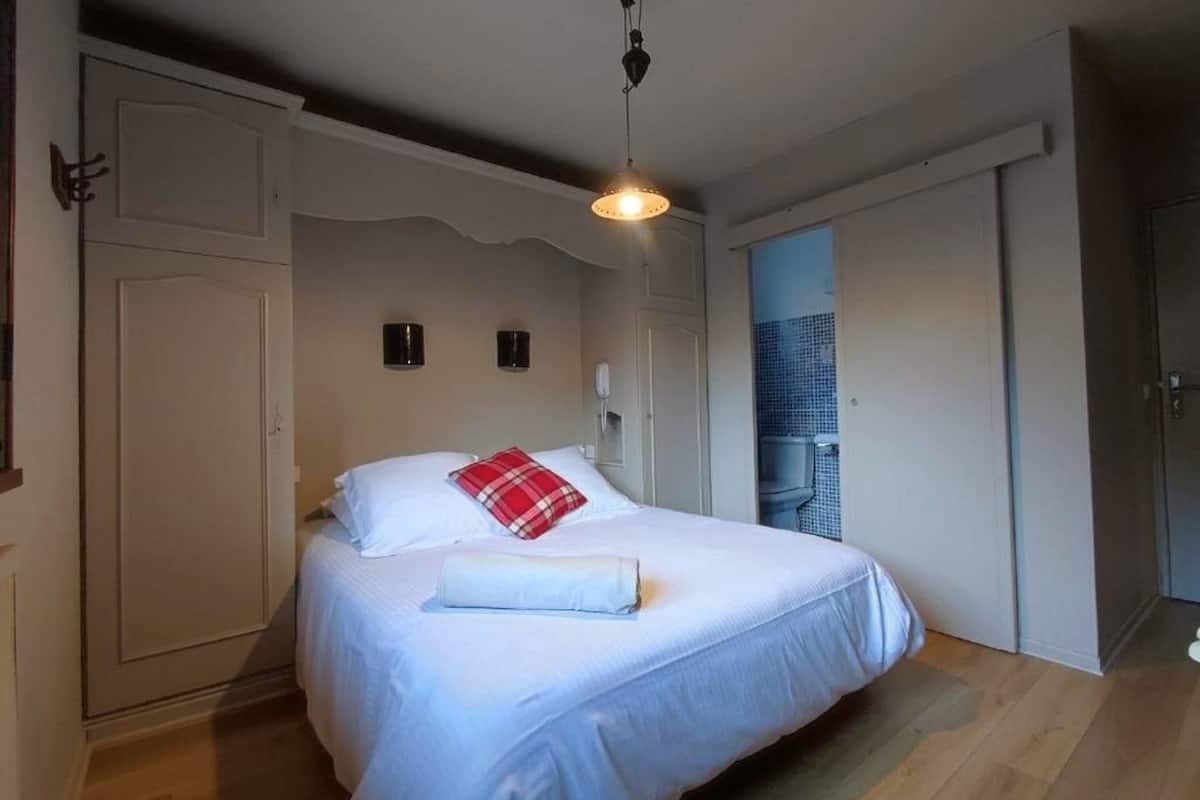 A bedroom with a bed, a wardrobe, a hanging light fixture, and a view of a cityscape through an open door.