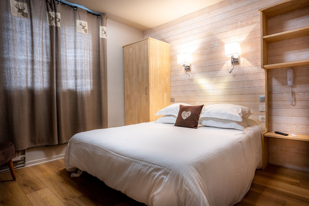 A neatly made bed with a heart-shaped pillow, wooden wardrobe, and a nightstand with a phone.