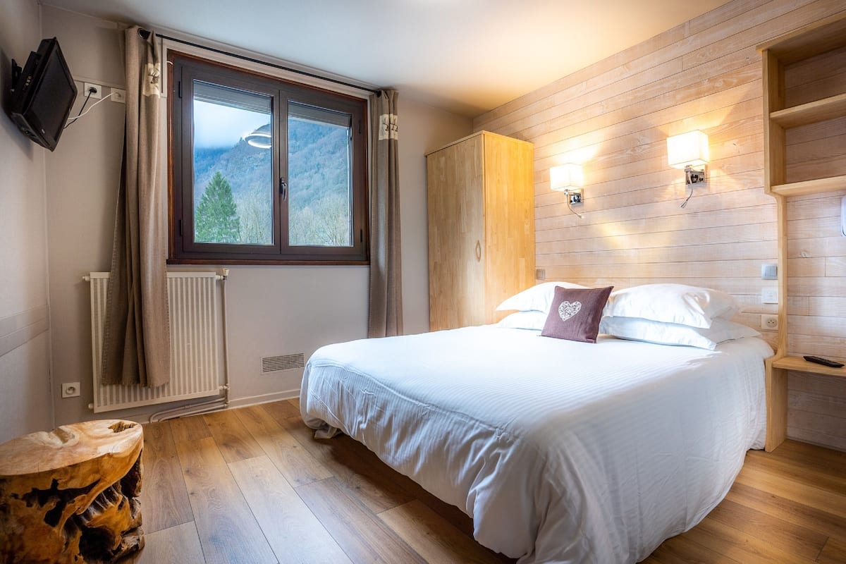 A bedroom with a large bed, wooden walls, a window with a mountain view, a radiator, and a wooden nightstand.