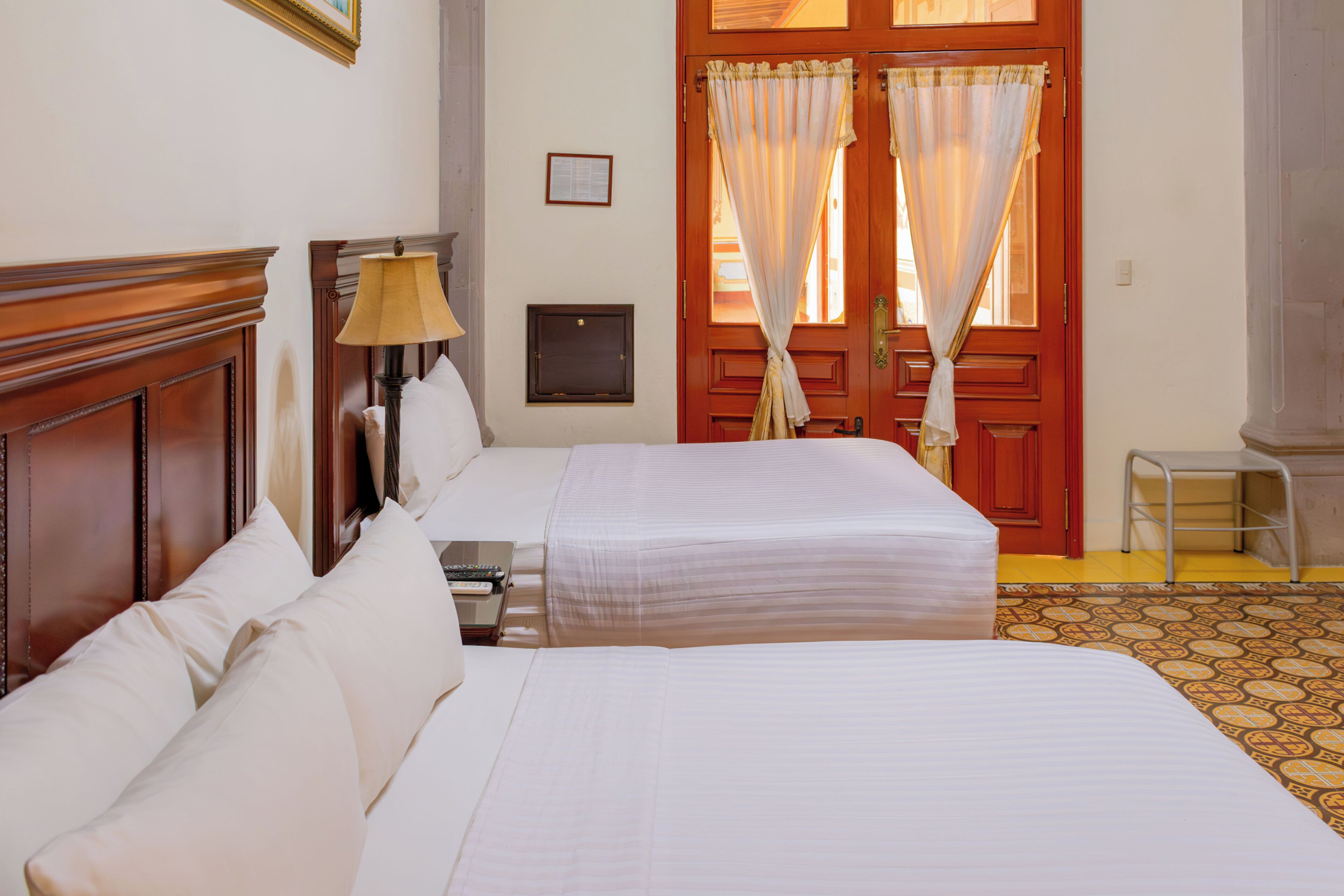 Habitación de hotel con dos camas, una mesita de noche, una lámpara, cabecera de madera, puerta con cortinas y un taburete pequeño.