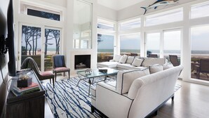 A spacious living room with a large sofa, a glass coffee table, and a fireplace. There are large windows offering a view of the ocean.