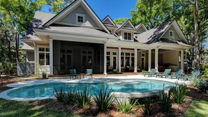 Una casa de dos pisos con una piscina grande, un porche cubierto con sillas y un jardín bien cuidado.