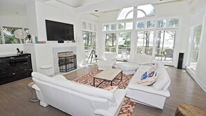 A spacious living room with white furniture, a fireplace, and large windows offering a view of the outdoors.