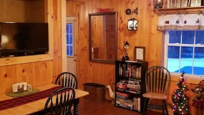 Una habitación acogedora con paredes de madera, un televisor montado en la pared, una estantería con juegos de mesa, un pequeño árbol de Navidad y una ventana con vistas al exterior.