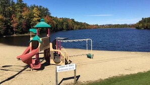 Un parque infantil con tobogán y estructura de escalada cerca de un lago, rodeado de árboles.