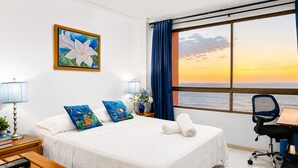 A hotel room with a bed, a desk, a chair, a window with a sunset view, a painting, and a lamp. - The best apartment - sea views (Santa Marta)