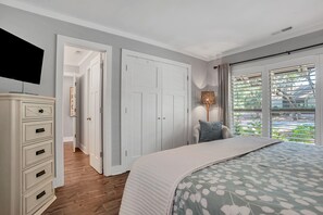 Une chambre à coucher comprenant un lit, une commode, un téléviseur, une porte et une fenêtre avec des stores.