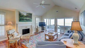 A spacious living room with a fireplace, a ceiling fan, a large TV, and a view of the outdoors through large windows. - 50 Lands End at The Sea Pines Resort (Hilton Head Island)