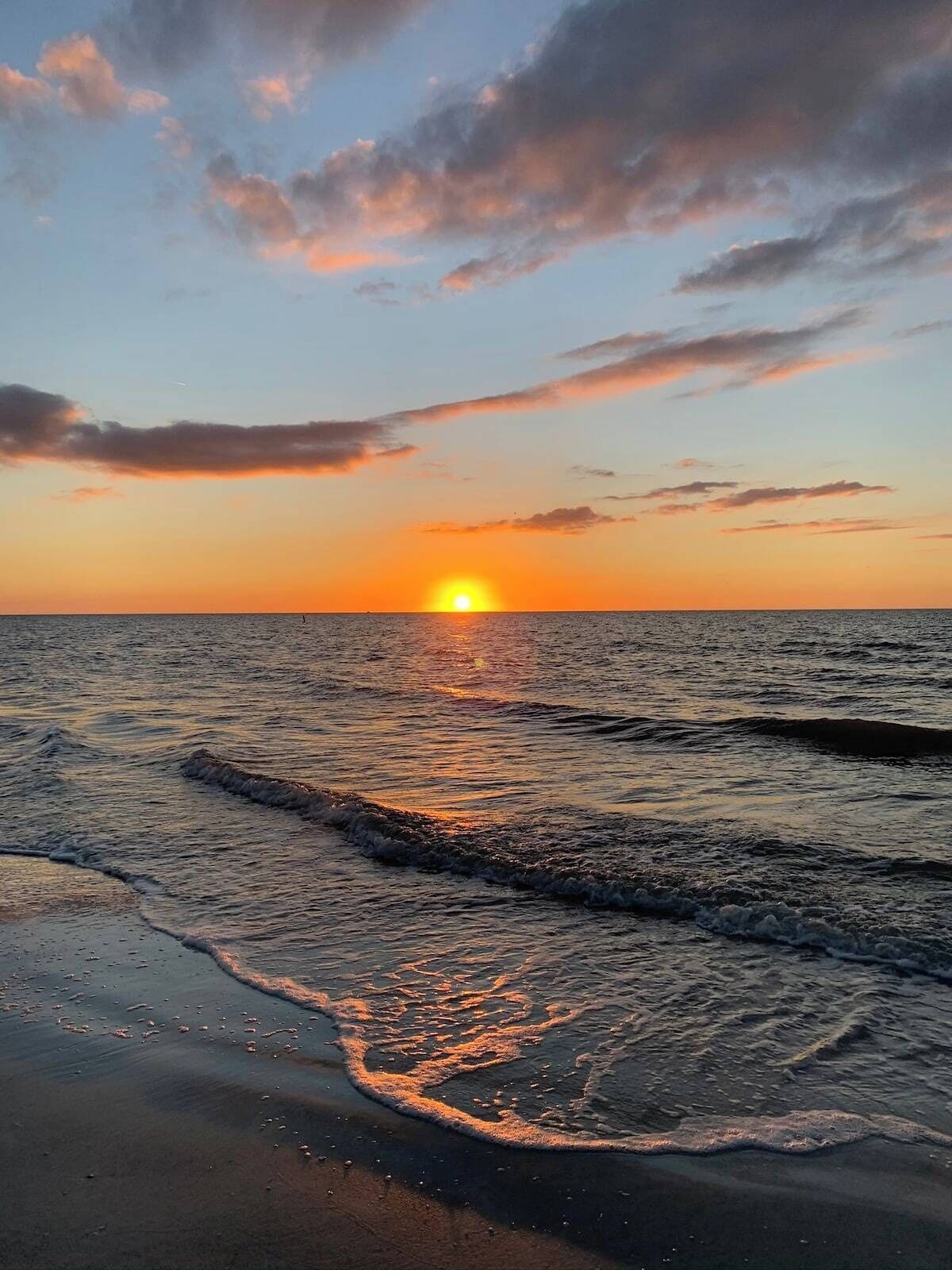 Un coucher de soleil sur une mer calme aux vagues douces, avec un horizon dégagé et quelques nuages dans le ciel.