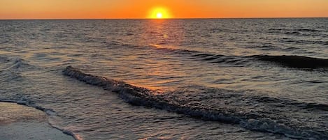 Un coucher de soleil sur une mer calme aux vagues douces, avec un horizon dégagé et quelques nuages dans le ciel.