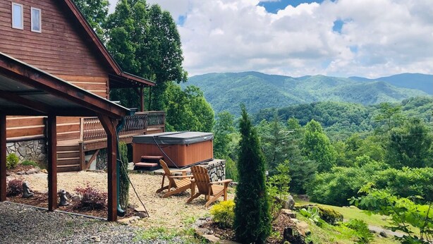 A wooden cabin with a hot tub and outdoor seating, overlooking a mountainous landscape.