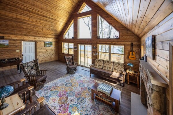 A cozy cabin interior with wooden walls, a large window, a fireplace, and a guitar.