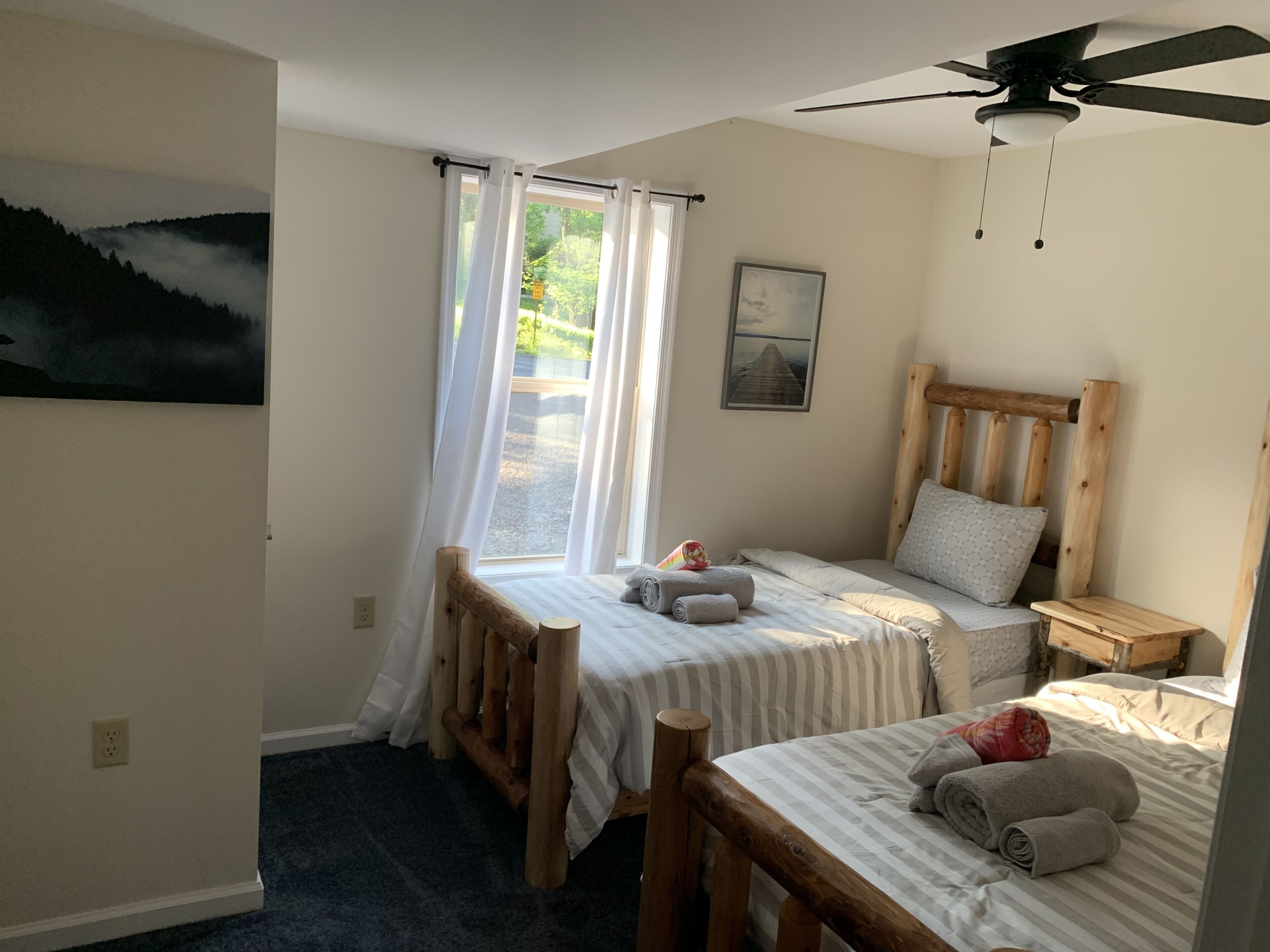 Un dormitorio con dos camas, un ventilador de techo, una ventana con cortinas y un cuadro en la pared.