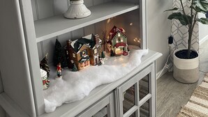 Een wit houten plankje met kaarsen, een decoratieve glazen vaas en kleine kersthuisjes.