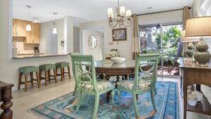 A dining area with a round table, green upholstered chairs, a chandelier, and a view of greenery outside.