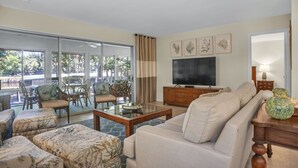 A spacious living room with a sofa, coffee table, and dining area with chairs. A flat-screen TV is mounted on the wall, and there are framed pictures above it.