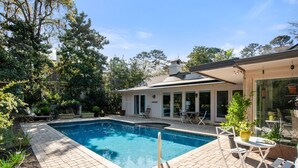A swimming pool with a tiled deck, surrounded by greenery and a house with a covered patio. - 337 Greenwood Dr. at The Sea Pines Resort (Hilton Head Island)