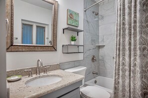 A bathroom with a sink, toilet, and bathtub. There are two shelves above the sink, one holding a plant and the other a small basket. A shower curtain with a paisley pattern is visible. - 536 Plantation Club Villas at The Sea Pines Resort (Hilton Head Island)