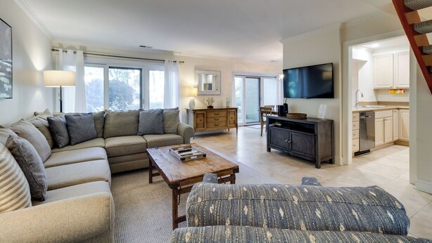 A spacious living room with a sofa, a coffee table, and a television. There is a kitchen area with cabinets and a staircase. - 540 Plantation Club Villas at The Sea Pines Resort (Hilton Head Island)