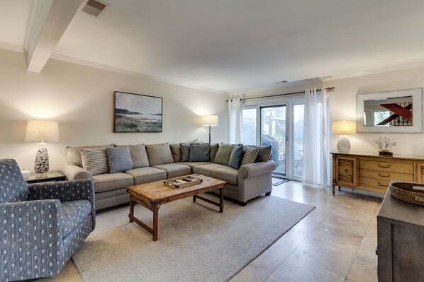 Una sala de estar con un sofá seccional, una mesa de centro y una mesita de noche con una lámpara. Hay una ventana con cortinas y vistas al exterior.