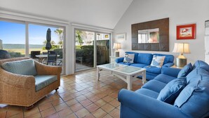 A living room with a blue sofa, wicker armchair, and a glass coffee table. There is a sliding glass door leading to an outdoor patio with a table and chairs. - 708 Schooner Court at The Sea Pines Resort (Hilton Head Island)