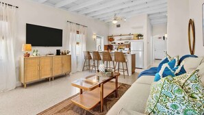 A living room with a sofa, a coffee table, and a TV. There is a kitchen in the background.