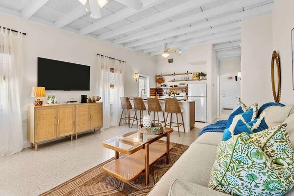 A living room with a sofa, a coffee table, and a TV. There is a kitchen in the background.