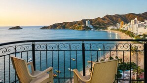 Un balcon avec deux fauteuils en osier, donnant sur une plage où l’on aperçoit des bateaux, un paysage urbain et des montagnes.